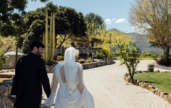 Pareja de recién casados caminando de la mano en un entorno natural rodeado de jardines, cactus y árboles bajo un cielo despejado en Casa Benigalip.